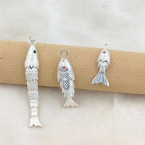 May include: Three silver-toned fish pendants with textured scale patterns. The longest pendant features a green eye, the middle pendant has an orange eye, and the smallest pendant has a red eye. Each piece includes a small loop for attachment.
