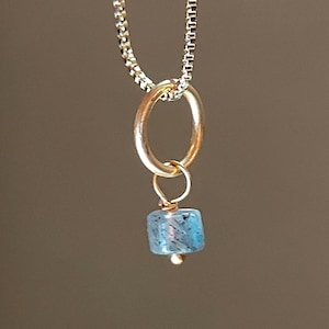 Peut inclure: Un collier à chaîne carrée dorée avec un anneau pendentif circulaire doré. Suspendue à l'anneau se trouve une petite perle bleue translucide de forme carrée avec des inclusions sombres, fixée par une fine boucle en fil doré et une petite perle.
