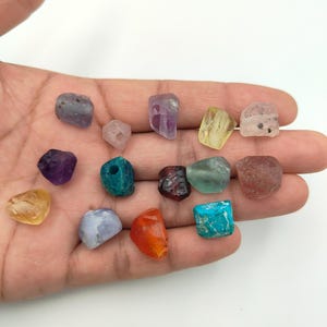 May include: A collection of fourteen small, irregular, tumbled gemstones in various colors including purple, blue, teal, orange, yellow, and clear. Each stone features a smooth, polished surface with unique natural textures and shapes.