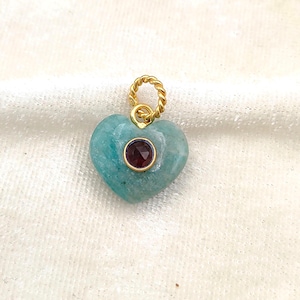 May include: A heart-shaped pendant crafted from smooth, light teal stone, featuring a small, round, dark red faceted gemstone set in a gold-toned bezel at the center, topped with a twisted gold-toned metal jump ring for attachment.