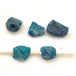 May include: Five raw, unpolished blue apatite gemstone beads with irregular, jagged shapes and natural textures. Three beads are positioned at the top, while two beads are threaded onto a thin wooden toothpick at the bottom, showing drilled holes.