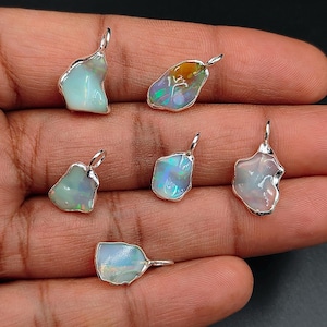 May include: Six irregular, raw opal pendants set in silver bezels with attached jump rings. Each gemstone displays a unique, milky white base with iridescent flashes of green, blue, and orange light, showcasing natural, unpolished stone textures.