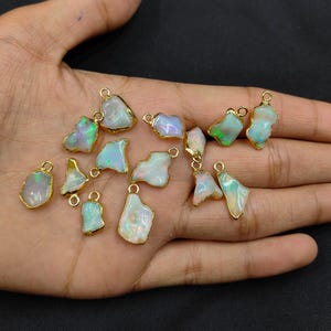 May include: A selection of opal pendants with gold-coloured edging. The opals are naturally shaped and show iridescent colours of green, blue, and white. Each pendant has a small loop for attaching to a chain or other jewellery component.