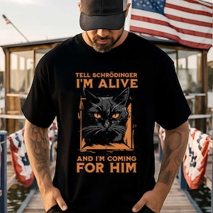 May include: A black short-sleeved t-shirt featuring a graphic of a black cat inside an orange square frame. The text reads "TELL SCHRÖDINGER I'M ALIVE AND I'M COMING FOR HIM" in orange capital letters above and below the cat illustration.