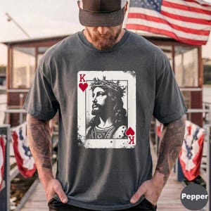 May include: A charcoal gray t-shirt featuring a graphic print of a playing card. The design shows a black and white illustration of a figure with a crown of thorns, framed by red heart symbols and the letter K in the top left and bottom right corners.