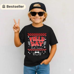 Può includere: T-shirt nera con le parole "FIELD DAY Red Team" in rosso, bianco e nero. Il design include motivi a scacchi, stelle e una faccina sorridente con occhiali da sole. Il bambino indossa un berretto nero e occhiali da sole.