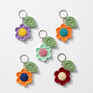 Puede incluir: Cinco llaveros de flores de ganchillo hechos a mano en varios colores: morado, naranja, verde, rojo y azul. Cada flor tiene un centro contrastante y una hoja verde, unidas a un llavero plateado. Los llaveros están hechos con un material texturizado similar al hilo.