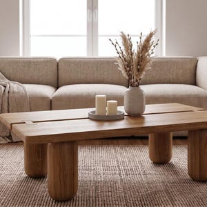 Op de afbeelding: Een rechthoekige houten salontafel met een glad blad en vier dikke, afgeronde cilindrische poten. De tafel heeft een zichtbaar verbindingsdetail op het oppervlak en staat voor een beige bank met een gedrapeerde plaid.