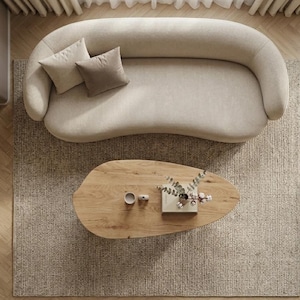 May include: A curved beige sofa with two throw pillows in cream and taupe, positioned above an organic-shaped wooden coffee table. The table holds a small white cup, a decorative object, and a vase with dried branches on a rectangular base.