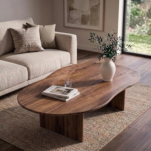 May include: A solid wood, oval-shaped coffee table with a natural finish. The table has a unique, organic shape and sits on a sturdy base. A white vase with greenery, a glass, and a book are on the table. The table is on a woven rug.