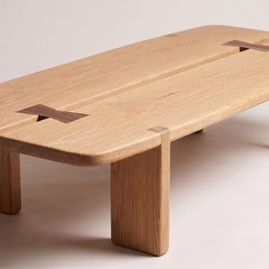 May include: A rectangular light-colored wooden coffee table with rounded corners and a central split down the length of the top. The table features two dark-colored butterfly-shaped wooden inlays connecting the split sections and four legs.