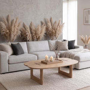 Puede incluir: Un sofá seccional gris claro con cojines beige y gris oscuro, junto a una mesa de centro de madera clara con una bandeja rectangular, dos velas cilíndricas y un jarrón de cristal con ramas, sobre una alfombra texturizada de color blanco roto.
