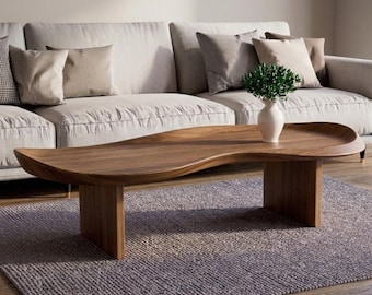 May include: A low-profile coffee table crafted from solid brown wood with a smooth, organic, kidney-bean-shaped top and two sturdy vertical slab legs. A white ceramic vase containing a small green plant sits on the table surface.