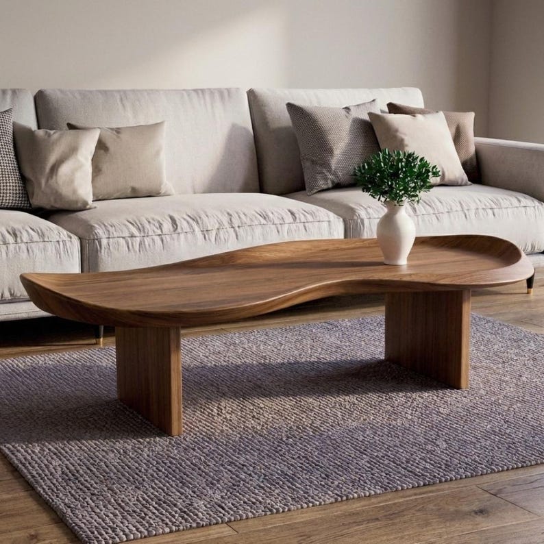 May include: A low-profile coffee table crafted from solid brown wood with a smooth, organic, kidney-bean-shaped top and two sturdy vertical slab legs. A white ceramic vase containing a small green plant sits on the table surface.