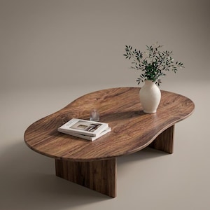 Pode incluir: Uma mesa de centro em forma de rim feita de madeira castanha escura com veios vis&iacute;veis. A mesa possui duas pernas verticais em painel. Na superf&iacute;cie, encontram-se um vaso de cer&acirc;mica branca com ramos verdes, dois livros e um copo.