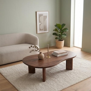 May include: A dark wood coffee table with an organic, kidney-bean shape and three thick, cylindrical legs. The table surface holds a small ceramic vase with a dried branch, a tall thin candle in a gold holder, and two stacked hardcover books.
