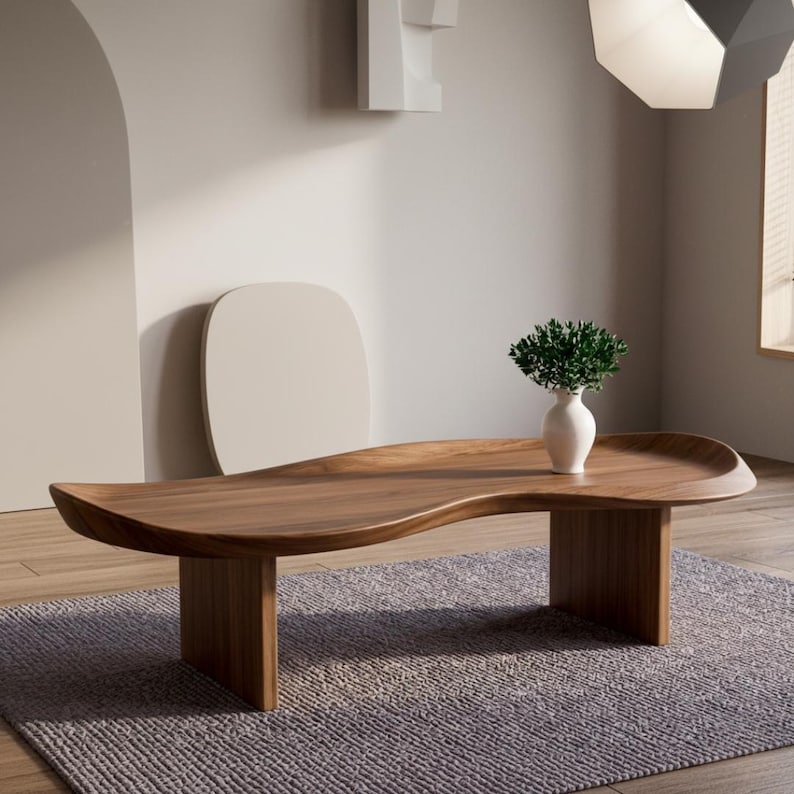May include: A low-profile, kidney-shaped coffee table crafted from rich, dark brown wood with a smooth, curved surface and two sturdy slab legs. A small, white ceramic vase containing a lush green plant sits on the table's surface.