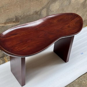 May include: A dark reddish-brown wooden bench featuring a smooth, kidney-shaped seat with a visible wood grain pattern. The seat is supported by two solid, rectangular vertical legs that match the deep, polished finish of the top surface.
