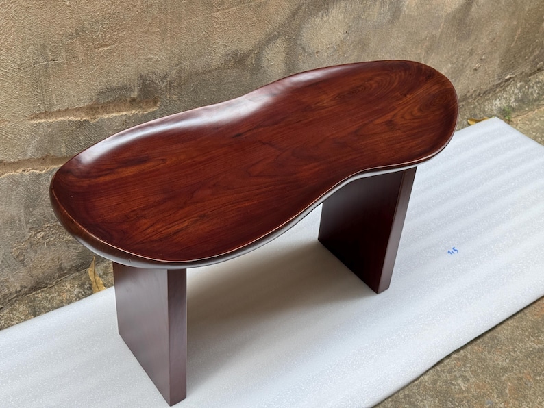 May include: A dark reddish-brown wooden bench featuring a smooth, kidney-shaped seat with a visible wood grain pattern. The seat is supported by two solid, rectangular vertical legs that match the deep, polished finish of the top surface.
