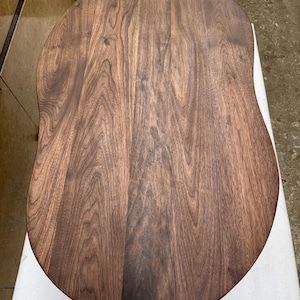 Pode incluir: Um tampo de mesa de madeira maci&ccedil;a com uma forma oval irregular e org&acirc;nica. A superf&iacute;cie apresenta uma cor castanha escura rica com padr&otilde;es de veios de madeira proeminentes e ondulados que correm verticalmente ao longo da t&aacute;bua polida.