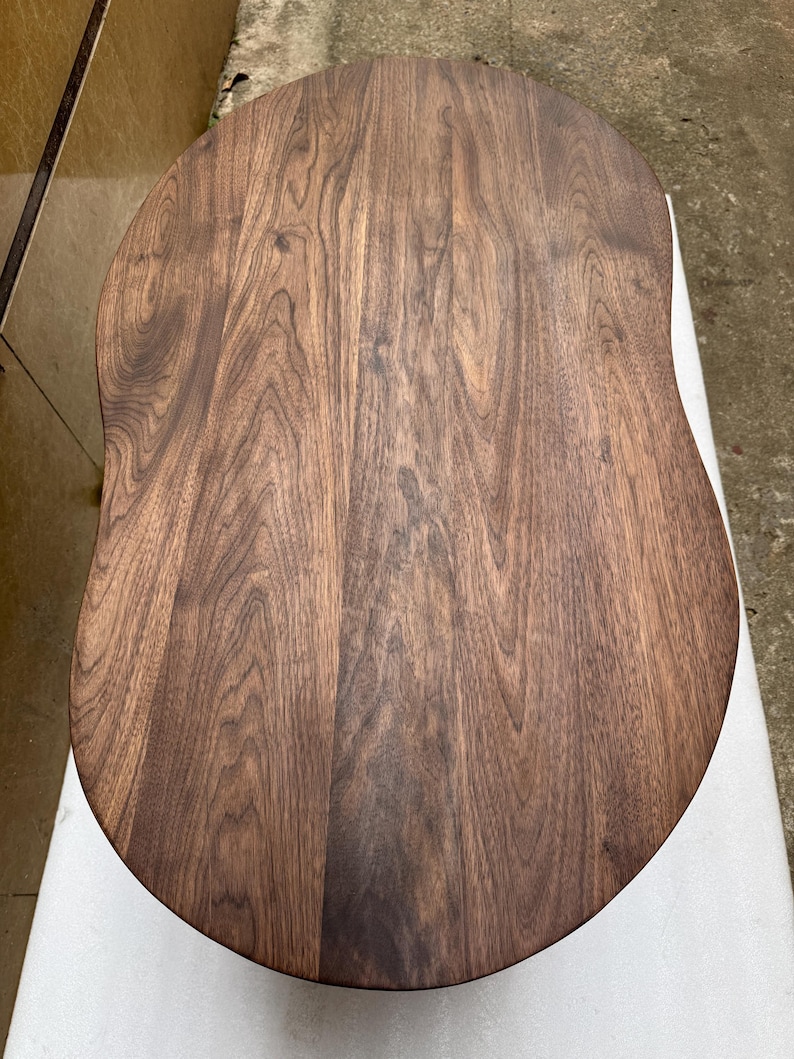 Pode incluir: Um tampo de mesa de madeira maci&ccedil;a com uma forma oval irregular e org&acirc;nica. A superf&iacute;cie apresenta uma cor castanha escura rica com padr&otilde;es de veios de madeira proeminentes e ondulados que correm verticalmente ao longo da t&aacute;bua polida.