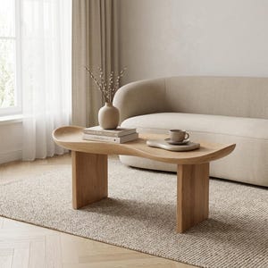 Puede incluir: Una mesa de centro de madera clara con un tablero de forma curva y orgánica y dos patas rectangulares robustas. Sobre la mesa hay un jarrón beige mate con ramas, dos libros apilados y una pequeña taza de cerámica sobre un plato irregular.