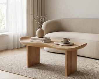 May include: A light wood coffee table with a curved, organic-shaped top and two sturdy rectangular legs. On the table sits a matte beige vase containing thin branches, two stacked books, and a small ceramic cup on a matching irregular saucer.