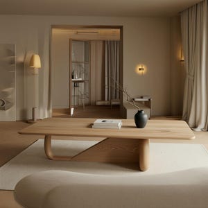Puede incluir: Una mesa de centro de madera clara con una superficie rectangular lisa y una base curva. Sobre la mesa hay un pequeño jarrón negro con una rama desnuda y dos libros blancos apilados. La mesa descansa sobre una alfombra de color claro.