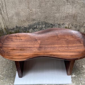May include: A low-profile coffee table crafted from dark brown wood with a smooth, polished finish. The tabletop features an irregular, organic kidney-bean shape with a slightly recessed center and raised edges, supported by two sturdy vertical legs.