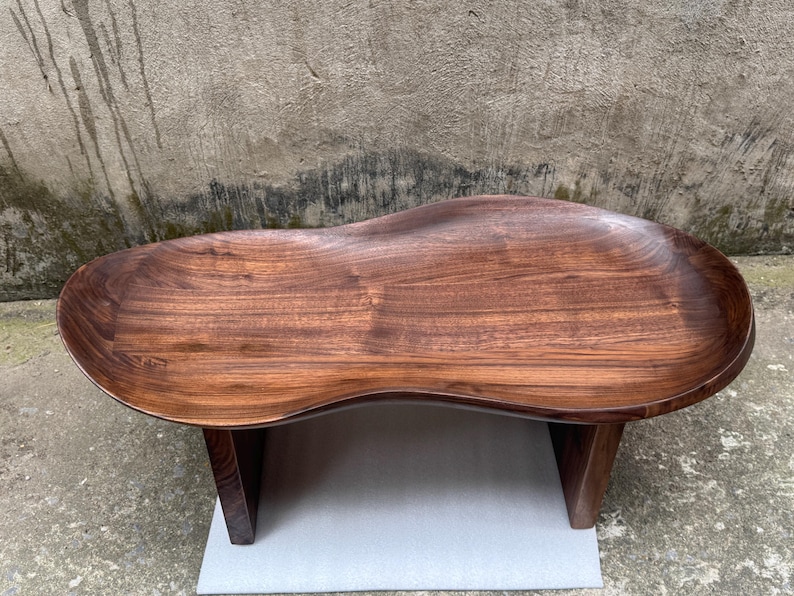 May include: A low-profile coffee table crafted from dark brown wood with a smooth, polished finish. The tabletop features an irregular, organic kidney-bean shape with a slightly recessed center and raised edges, supported by two sturdy vertical legs.