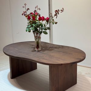 Pode incluir: Uma mesa de centro de madeira castanho-escura com um tampo org&acirc;nico em forma de rim e duas pernas verticais planas e robustas. Um vaso de vidro transparente canelado com um ramo de rosas vermelhas e folhas verdes est&aacute; no centro da mesa.