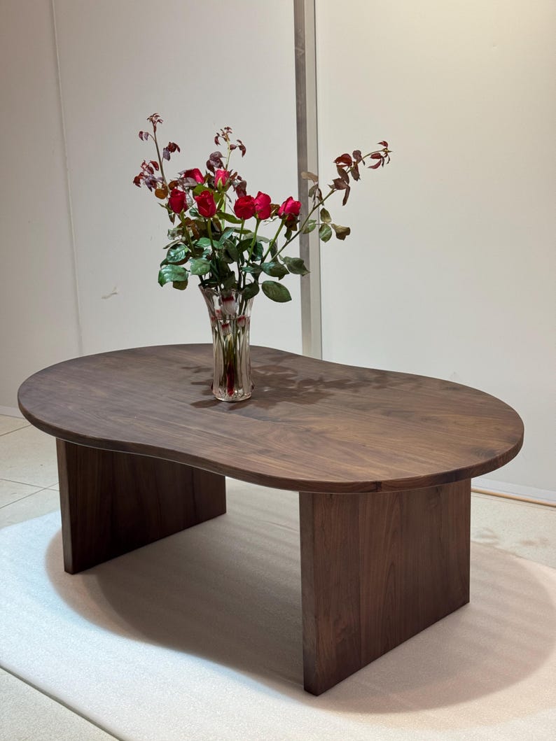 Pode incluir: Uma mesa de centro de madeira castanho-escura com um tampo org&acirc;nico em forma de rim e duas pernas verticais planas e robustas. Um vaso de vidro transparente canelado com um ramo de rosas vermelhas e folhas verdes est&aacute; no centro da mesa.