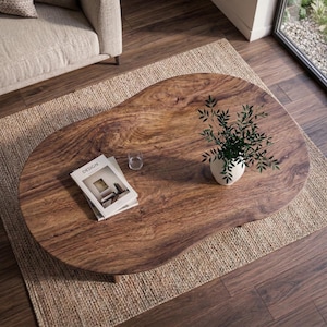 Pode incluir: Uma mesa de centro em forma de rim feita de madeira castanha maci&ccedil;a com um padr&atilde;o de gr&atilde;o natural vis&iacute;vel. Na superf&iacute;cie, encontra-se um vaso de cer&acirc;mica branca com uma planta verde, um copo transparente e dois livros com a palavra DESIGN.