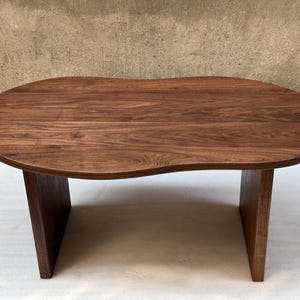 Pode incluir: Uma mesa de centro de madeira castanho-escura com um tampo org&acirc;nico em forma de rim, apresentando uma superf&iacute;cie lisa e polida que exibe padr&otilde;es naturais de gr&atilde;o de madeira. A mesa &eacute; suportada por duas pernas verticais s&oacute;lidas.
