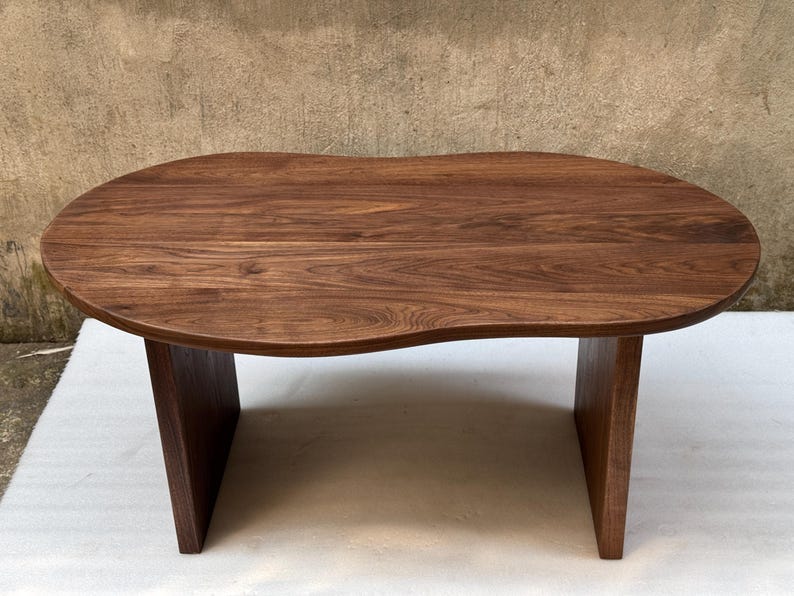 Pode incluir: Uma mesa de centro de madeira castanho-escura com um tampo org&acirc;nico em forma de rim, apresentando uma superf&iacute;cie lisa e polida que exibe padr&otilde;es naturais de gr&atilde;o de madeira. A mesa &eacute; suportada por duas pernas verticais s&oacute;lidas.