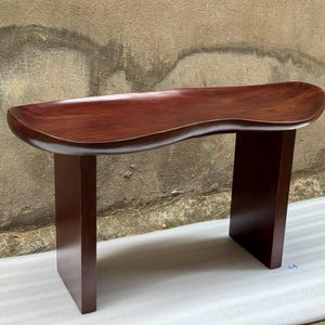 May include: A dark brown wooden bench featuring a smooth, curved, kidney-shaped seat surface with a polished finish. The bench is supported by two vertical, rectangular slab legs that extend from the underside of the seat to the floor.