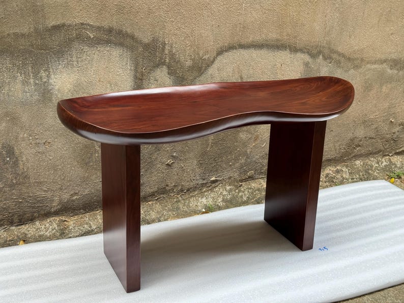 May include: A dark brown wooden bench featuring a smooth, curved, kidney-shaped seat surface with a polished finish. The bench is supported by two vertical, rectangular slab legs that extend from the underside of the seat to the floor.