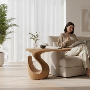 Puede incluir: Una mesa auxiliar de madera clara con una base curva en forma de S y una superficie superior plana. La mesa está colocada junto a un sofá beige y una palmera en maceta en una habitación luminosa con suelo claro y cortinas blancas.