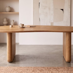 Puede incluir: Una mesa de comedor de madera de color marrón claro con un tablero orgánico de forma asimétrica y dos patas gruesas y redondeadas. La superficie es lisa y presenta un patrón de veta de madera natural, sosteniendo una taza de cerámica.