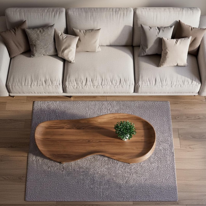 May include: A beige three-cushion sofa with various brown and beige throw pillows sits above a wooden coffee table with an irregular, organic shape. A small green potted plant rests on the table, which is placed on a textured gray area rug.