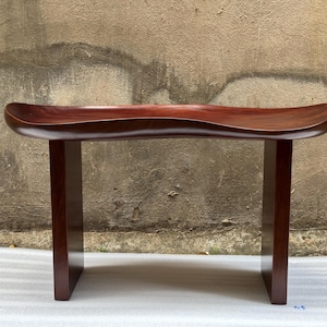 May include: A dark reddish-brown wooden bench featuring a smooth, contoured, saddle-shaped seat supported by two vertical rectangular legs. The wood grain is visible across the polished surface, which curves upward at both ends of the seat.