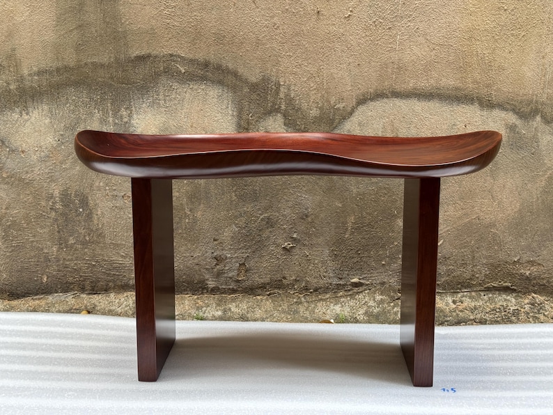 May include: A dark reddish-brown wooden bench featuring a smooth, contoured, saddle-shaped seat supported by two vertical rectangular legs. The wood grain is visible across the polished surface, which curves upward at both ends of the seat.