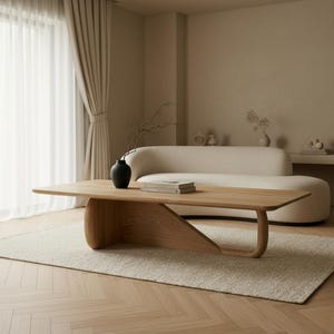 Puede incluir: Una mesa de centro de perfil bajo fabricada en madera de tono claro, con una superficie rectangular lisa y una base escultural única con una estructura de soporte curva y continua. La mesa sostiene un jarrón negro con ramas y libros.