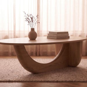 May include: An oval light wood coffee table with a curved, sculptural base. On the tabletop sits a small, rounded beige vase containing dried stems and a stack of two books. The table features a smooth, natural wood grain finish throughout.