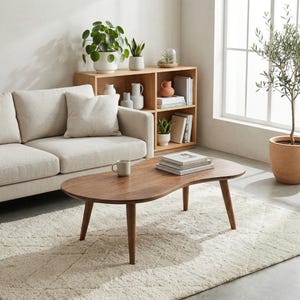 May include: A kidney-shaped coffee table made of dark brown wood with three tapered legs. The table surface holds a light gray ceramic mug and a stack of three books. The table is positioned in front of a light beige sofa on a textured rug.