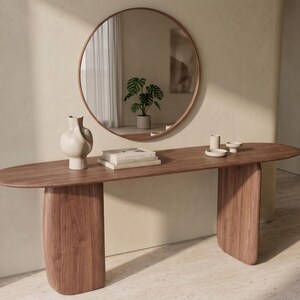 May include: A long, oval-shaped wooden console table with two thick, curved slab legs. On the table sits a tall, off-white ceramic vase, a stack of two books, and two small ceramic dishes. A large, round, thin-framed mirror hangs above.