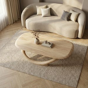 May include: A light wood coffee table with an oval top and a curved, crescent-shaped base. The table holds a small ceramic vase with dried branches and a stack of two books. The furniture is positioned on a textured rug in front of a curved sofa.