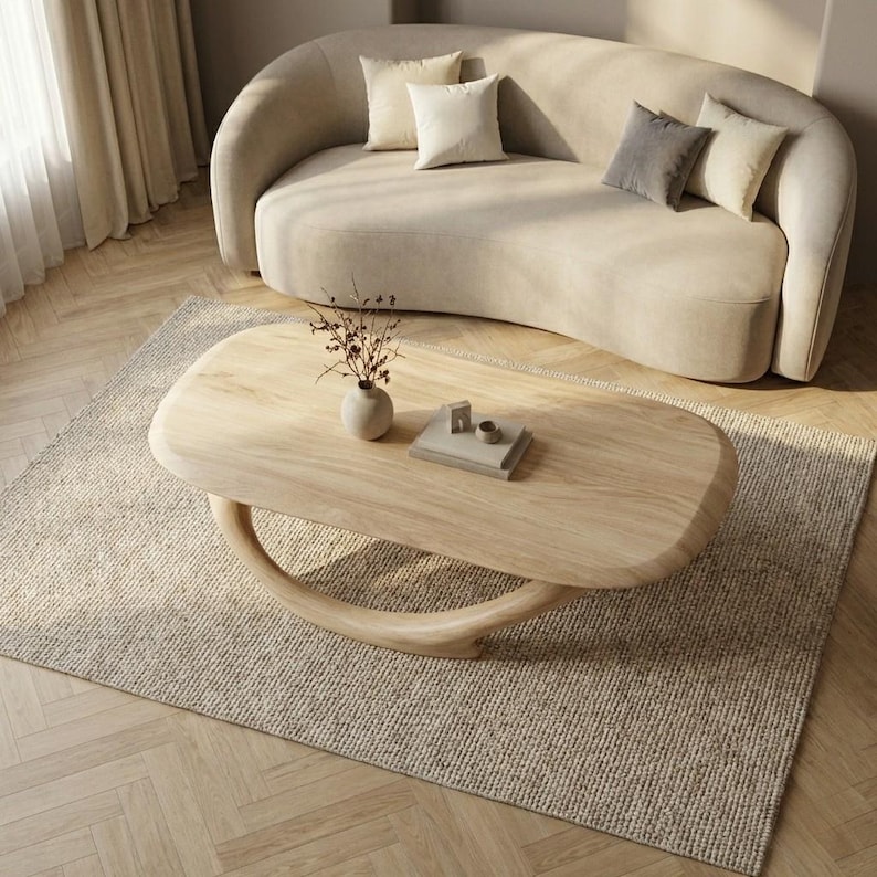May include: A light wood coffee table with an oval top and a curved, crescent-shaped base. The table holds a small ceramic vase with dried branches and a stack of two books. The furniture is positioned on a textured rug in front of a curved sofa.