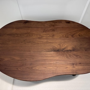 Pode incluir: Uma mesa de centro de madeira castanho-escura com um tampo liso de forma org&acirc;nica semelhante a um feij&atilde;o. A superf&iacute;cie apresenta um padr&atilde;o de gr&atilde;o de madeira natural proeminente com estrias e n&oacute;s castanhos escuros, real&ccedil;ando a textura.