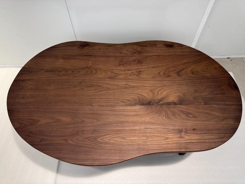 Pode incluir: Uma mesa de centro de madeira castanho-escura com um tampo liso de forma org&acirc;nica semelhante a um feij&atilde;o. A superf&iacute;cie apresenta um padr&atilde;o de gr&atilde;o de madeira natural proeminente com estrias e n&oacute;s castanhos escuros, real&ccedil;ando a textura.